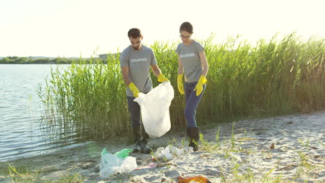 Couple-Of-Volunteers-Picking-Up-Trash-And-Collecting-It-In-The-Plastic-Bag-While-Cleaning-Lake-Beach