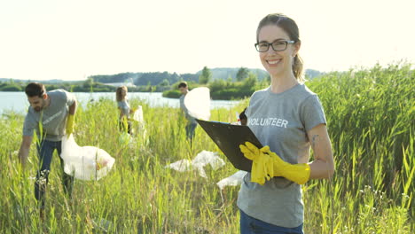 Young-Pretty-Woman-Standing-With-Alist-Of-Volunteers-In-Front-Of-The-Camera-And-Smiling-While-Students-Volunteering-Behind-And-Cleaning-Up-A-Lake-Area