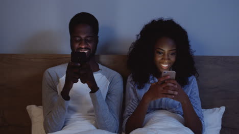 American-Couple-Laughing-While-Looking-At-Their-Cell-Phones-In-Bed-Before-Bedtime