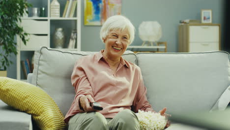 Portrait-Shot-Of-The-Old-Woman-Sitting-On-The-Couch-In-The-Living-Room,-Turning-On-Tv-With-A-Remote-Control-And-Laughing-While-Watching-Tv
