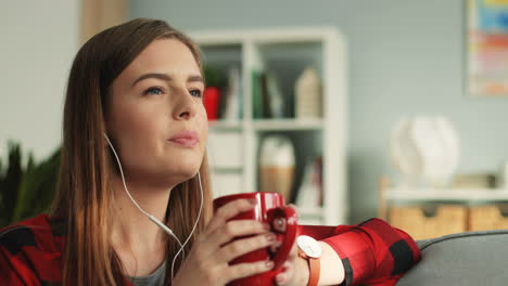 Close-Up-Of-The-Young-Happy-Beautiful-Girl-In-Headphones-Sipping-Hot-Tea-Or-Coffee,-Smiling-To-The-Camera-And-Listening-To-The-Music