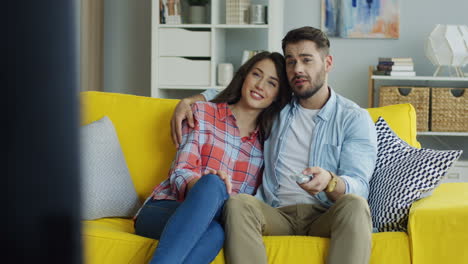 Portrait-Shot-Of-The-Good-Looking-Young-Man-And-Woman-Watching-Tv-Together-While-Resting-On-The-Yellow-Couch-In-The-Living-Room