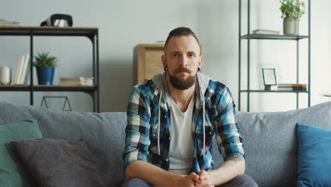 Serious-Upset-Hipster-Man-Sitting-On-The-Couch-In-The-Living-Room,-Looking-To-The-Camera-And-Then-Down