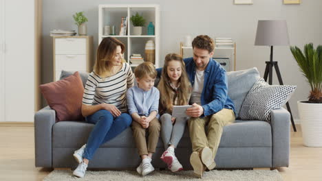 Joyful-Family-With-Two-Kids-Sitting-On-The-Sofa,-Smiling-While-Watching-Something-On-The-Tablet-Computer
