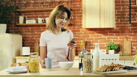 Pretty-Young-Woman-Having-Breakfast-In-The-Morning-In-The-Kitchen-And-Chatting,-Scrolling-And-Tapping-On-The-Smartphone
