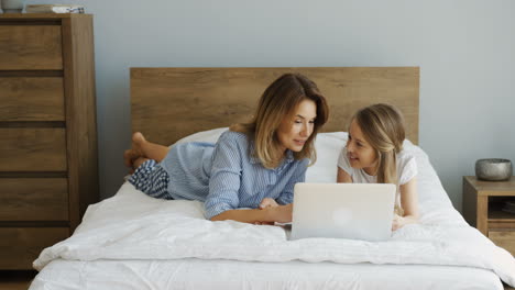 Cheerful-Smiled-Mother-And-Litle-Daughter-Resting-After-Waking-Up-In-The-Morning-On-The-Unmade-Bed-In-Front-Of-The-Laptop-Screen