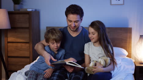 Handsome-Father-Reading-A-Fairytale-Book-For-His-Small-Son-And-Daughter-Before-They-Going-To-Sleep-In-The-Bedroom-At-Night