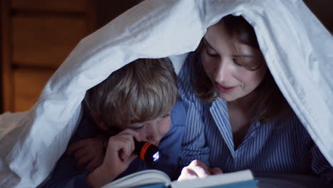 Cute-Small-Boy-And-His-Pretty-Mother-With-A-Little-Lantern-Reading-An-Interesting-Book-Under-The-Blanket-At-Night