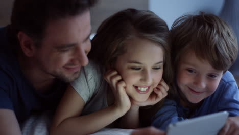 Close-Up-Of-The-Smiled-Father-And-His-Little-Son-And-Daughter-Lying-On-Their-Stomachs-And-Watching-Something-On-The-Tablet-Device-While-Dad-Telling-Or-Showing-Them-Something