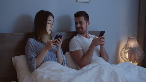Young-Attractive-Just-Married-Man-And-Woman-Sitting-In-Their-Bed-Late-At-Night-And-Playing-On-Their-Smartphones-While-Taping,-Then-Looking-At-Each-Other