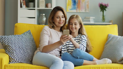 Schöne-Blonde-Mutter-Und-Hübsches-Jugendliches-Mädchen,-Das-Auf-Dem-Gelben-Sofa-Sitzt-Und-Etwas-Auf-Dem-Smartphone-Anschaut