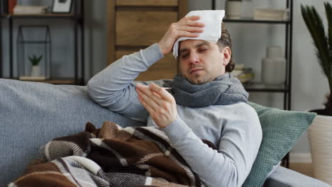 Unhealthy-Man-Covered-With-A-Plaid-Sitting-On-The-Gray-Sofa-At-Home,-Checking-His-Temperature-With-A-Thermometer-As-He-Having-An-Influenza-And-Fever,-Holding-A-Towel-On-The-Forehead