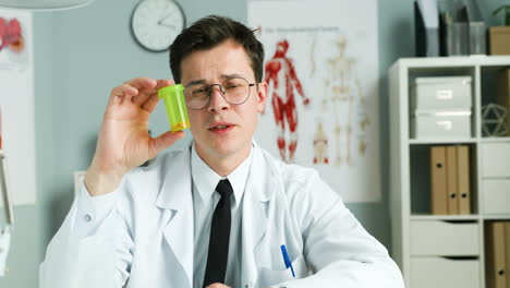 Young-Man-Medic-In-White-Gown-And-Glasses-Sitting-At-Table-In-Cabinet,-Showing-Pills-And-Remedy-To-Camera