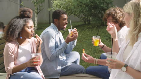 Group-Of-Multiethnic-Friends-Spending-Time-Together,-Eating-Nuggets-And-Drinking-Refreshing-Juice,-Sitting-In-A-Wooden-Bench-In-A-Park,-Talking-To-Each-Other-And-Having-Fun
