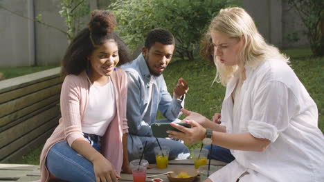 Young-Blonde-Woman-Showing-Something-Interesting-On-Mobile-Phone-To-Her-Multiethnic-Friends-In-A-Park
