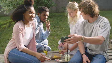 Young-Man-Showing-Something-Interesting-On-Mobile-Phone-To-His-Multiethnic-Friends-In-A-Park