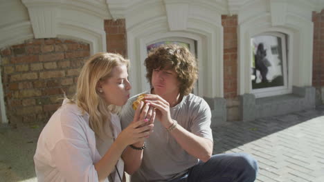 Young-Couple-Sharing-A-Tasty-Hambuger,-Sitting-On-The-Sidewalk-In-The-City