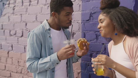 Young-Couple-Sharing-A-Tasty-Hamburger-And-Drinking-A-Cold-Drink-Together,-While-Laughing-Leaning-Against-A-Colourful-Brick-Wall