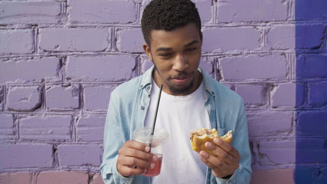 Happy-Young-Man-Eating-A-Delicious-Hamburger-And-Holding-A-Cold-Drink-In-Plastic-Cup,-Leaning-Against-A-Colorful-Brick-Wall-In-The-City