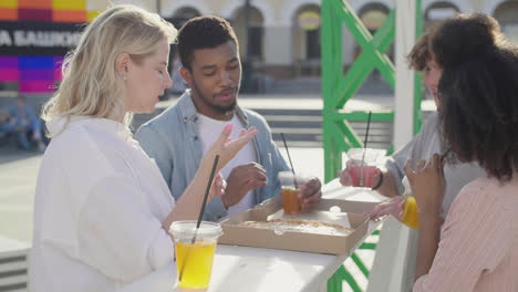 Blonde-Girl-Bringing-Pizza-To-Share-With-Her-Friends-While-Standing-At-An-Outdoor-Table-In-The-Street,-Drinking-Cold-Drinks-And-Chatting-Together