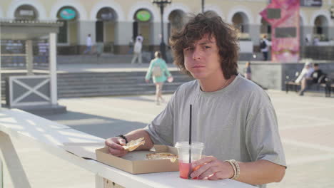 Young-Man-Eating-Pizza-And-Drinking-A-Cold-Drink-Alone-While-Standing-At-An-Outdoor-Table-In-The-Street