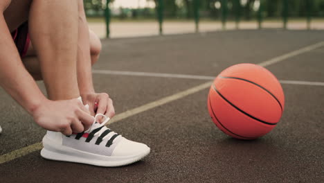 Close-Up-Of-A-Male-Basketball-Player-Putting-On-His-Shoe,-Tying-Shoeslaces-And-Then-Picking-The-Ball-Up-From-The-Floor-In-An-Outdoor-Basketball-Court