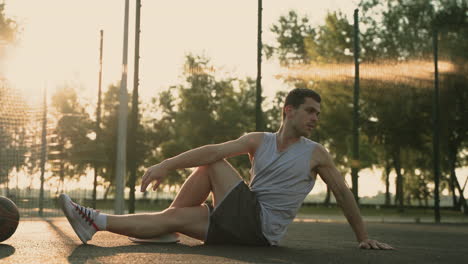 Concentrated-Male-Basketball-Player-Stretching-And-Doing-Seated-Spinal-Twist,-Sitting-On-Floor-In-Outdoor-Basketball-Court