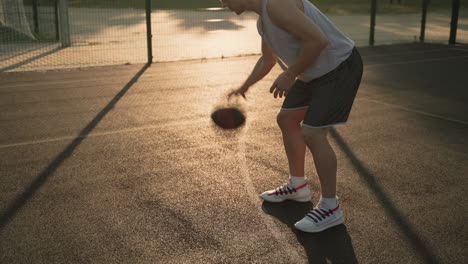 A-Male-Basketball-Player-Bouncing-And-Dribbling-The-Ball-Between-His-Legs-In-An-Outdoor-Court-At-Sunset