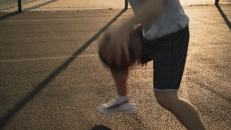 Nahaufnahme-Eines-Männlichen-Basketballspielers,-Der-Bei-Sonnenuntergang-Auf-Einem-Außenplatz-Den-Ball-Zwischen-Seinen-Beinen-Hüpft-Und-Dribbelt