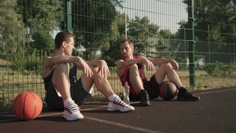Two-Happy-Basketball-Players-Chatting-While-Sitting-And-Leaning-Against-A-Metal-Fence-In-An-Outdoor-Basketball-Court