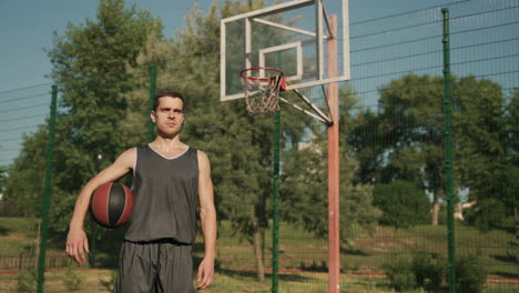 Concentrated-Handsome-Basketball-Player-Standing-And-Holding-The-Ball-In-An-Outdoor-Basketball-Court-In-A-Sunny-Day,-Looking-Ahead-Of-Him-With-Focused-And-Determined-Expression