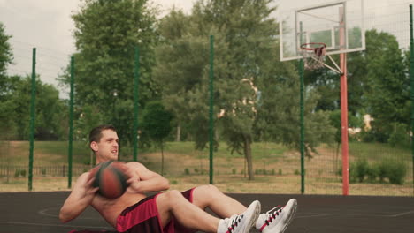 Handsome-Shirtless-Sportsman-Doing-Abdominal-Exercises,-Holding-A-Basketball-Ball-And-Lying-At-Ground-In-An-Outdoor-Basketball-Court