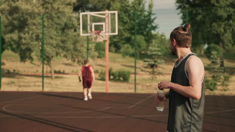 In-Foreground,-A-Handsome-Blonde-Bearded-Basketball-Player-Taking-A-Break-And-Drinking-Water-In-An-Outdoor-Basketball-Court