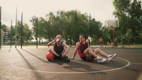 Two-Basketball-Players-Sitting-In-The-Center-Of-An-Outdoor-Basketball-Court,-Taking-A-Break,-Drinking-Water-And-Talking-To-Each-Other