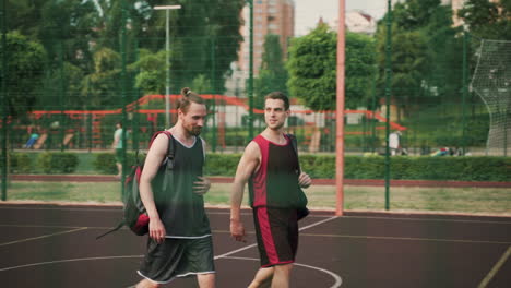 Two-Smiling-Handsome-Sportsmen-Walking-And-Talking-To-Each-Other,-While-Leaving-Together-The-Outdoor-Basketball-Court-After-Training-Session