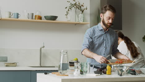A-Concentrated-Bearded-Man-Holding-Chopping-Board-While-Adding-Sliced-Tomatoes-In-Glass-Bowl-In-A-Modern-Kitchen