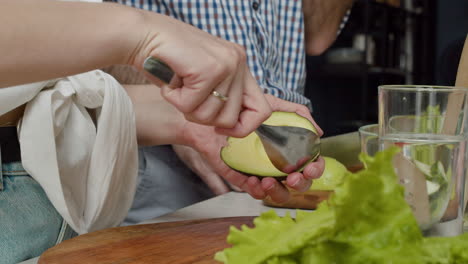 Close-Up-Of-A-Woman-Hands-Scooping-The-Flesh-Out-From-Avocado,-While-Man-Slicing-Avocado-In-A-Wooden-Chopping-Board-In-A-Modern-Kitchen