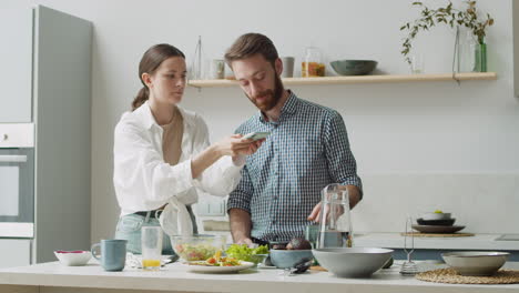 Woman-Taking-Photo-Of-Food-On-Table-At-Home,-While-Man-Watching-And-Telling-Something-Funny