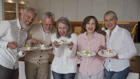 Group-Of-Happy-Senior-Friends-Having-Fun-Shaking-Plates-With-Flans,-While-Standing-And-Looking-At-Camera-In-A-Kitchen