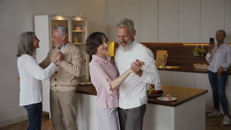 Two-Happy-Senior-Couples-Dancing-In-The-Kitchen,-While-A-Pretty-Elderly-Woman-Filming-Them-On-Mobile-Phone