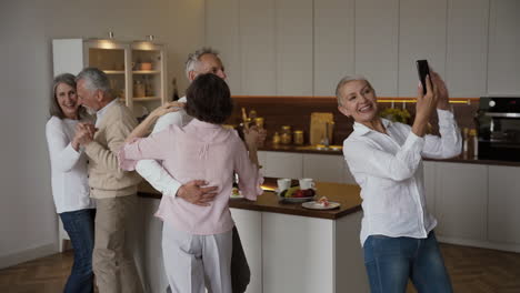Zwei-Glückliche-Seniorenpaare-Tanzen-In-Der-Küche,-Während-Eine-Hübsche-ältere-Frau-Ein-Selfie-Video-Mit-Dem-Handy-Macht