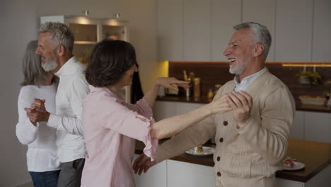 Two-Happy-Senior-Couples-Dancing-In-The-Kitchen,-While-On-Blurred-Background-A-Pretty-Elderly-Woman-Filming-Them-On-Mobile-Phone