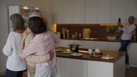 Two-Happy-Senior-Couples-Dancing-In-The-Kitchen,-While-On-Blurred-Background-A-Pretty-Elderly-Woman-Filming-Them-On-Mobile-Phone-1