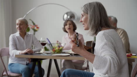 In-Foreground,-Happy-Senior-Woman-Singing-And-Playing-The-Guitar-Sitting-On-Chair,-While-In-Blurred-Background-Three-Elderly-Friends-Listening-To-Her-And-Singing-Together-Sitting-At-The-Table