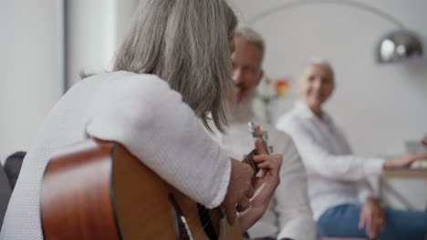 Happy-Senior-Woman-Singing-And-Playing-The-Guitar-Sitting-On-Chair,-While-In-Blurred-Background-Elderly-Friends-Listening-To-Her-And-Singing-Together-Sitting-At-The-Table