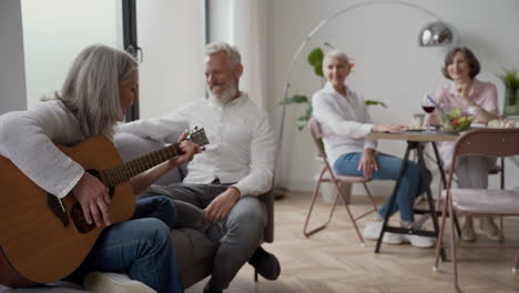 Happy-Senior-Woman-Singing-And-Playing-The-Guitar-Sitting-On-Chair,-While-In-Blurred-Background-Elderly-Friends-Listening-To-Her-And-Singing-Together-Sitting-At-The-Table-1