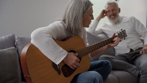Happy-Senior-Woman-Singing-And-Playing-The-Guitar-Sitting-On-Chair,-While-In-Blurred-Background-Elderly-Friends-Listening-To-Her-And-Singing-Together-Sitting-At-The-Table-2