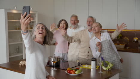 Happy-Senior-Woman-Making-A-Selfie-With-A-Smartphone-In-The-Kitchen,-Her-Friends-Laugh-And-Wave-At-The-Camera