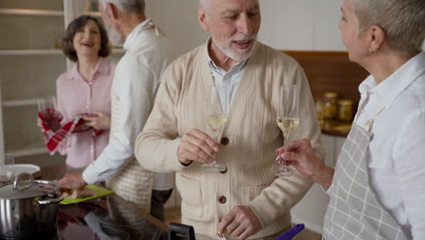 Older-Man-And-Woman-Friends-Toasting-With-A-Glass-Of-Wine,-While-A-Couple-Of-Older-Friends-Cooking-In-The-Kitchen-In-The-Background