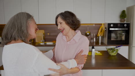 Senior-Woman-Waving-And-Hugging-Her-Older-Friend-In-The-Kitchen